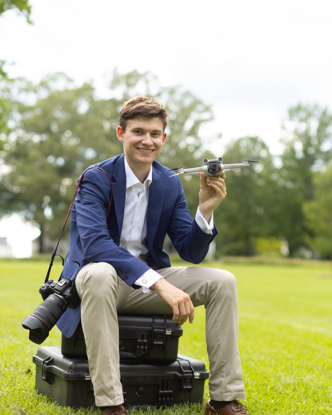 Samuel Long, who leads SLP Media, sitting on hard camera cases on a lawn with a Canon camera at his side and a small drone in his hand